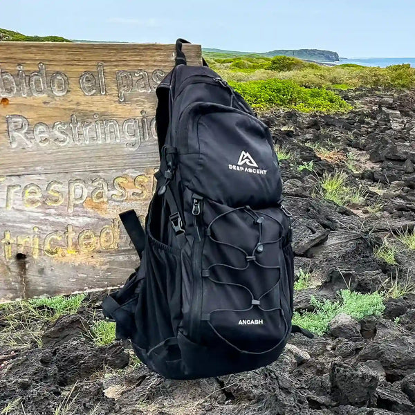 Ancash Packable Daypack hanging on a sign in a rocky field with the ocean in the background in San Cristobal, Galapagos.