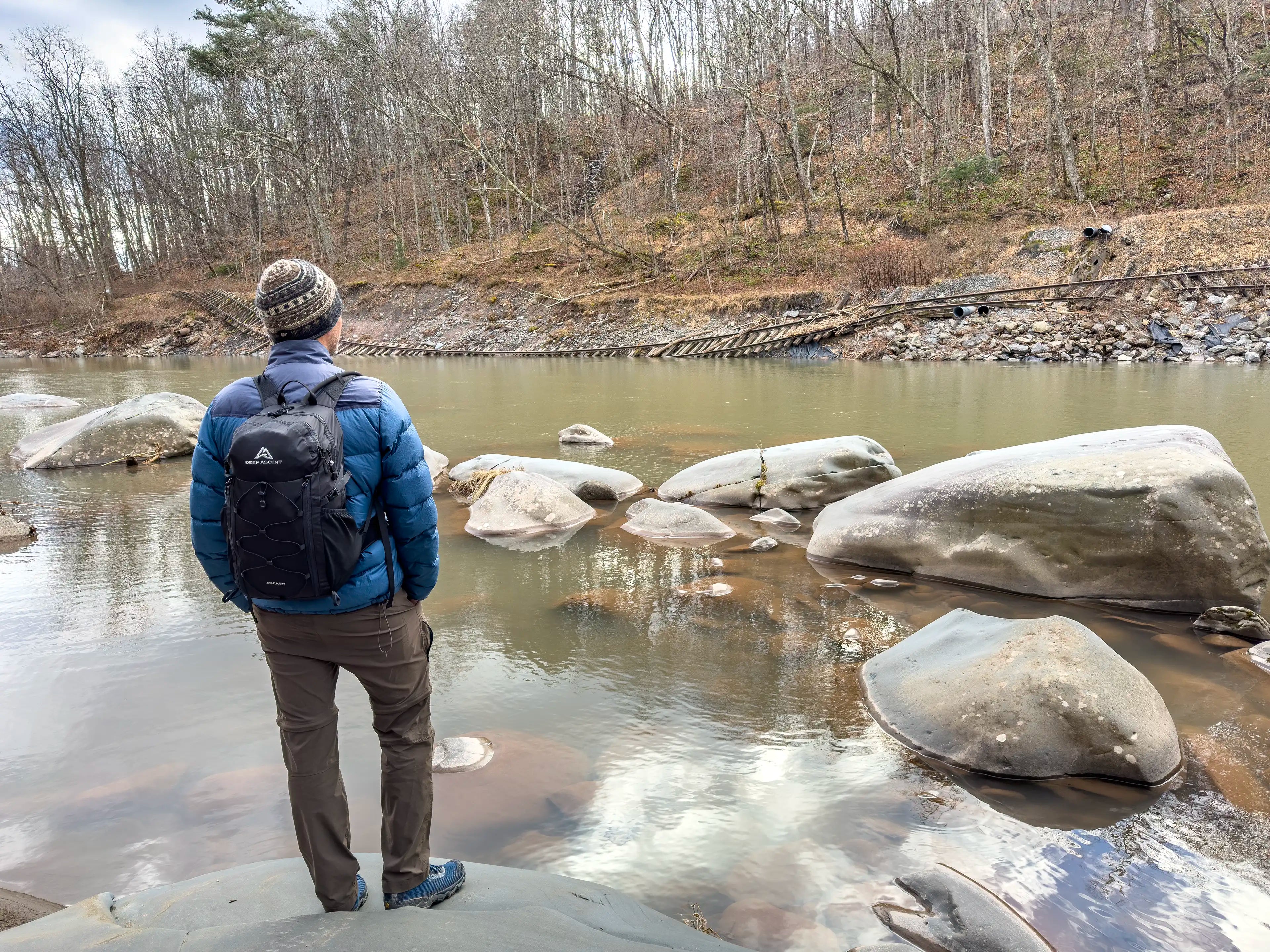 A male hiker standing on a rock next to the Esopus Creek in New York wearing the Ancash Packable Daypack by Deep Ascent.