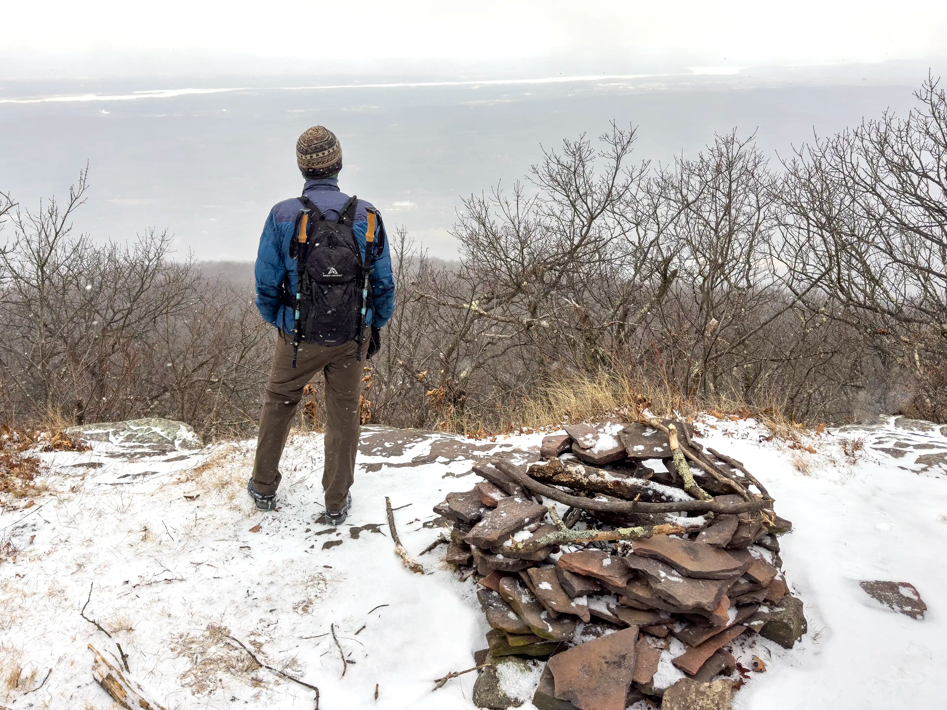 A male hiker standing in the snow next to a fire pit at an overlook on Overlook Mountain in New York wearing the Ancash Packable Daypack by Deep Ascent.