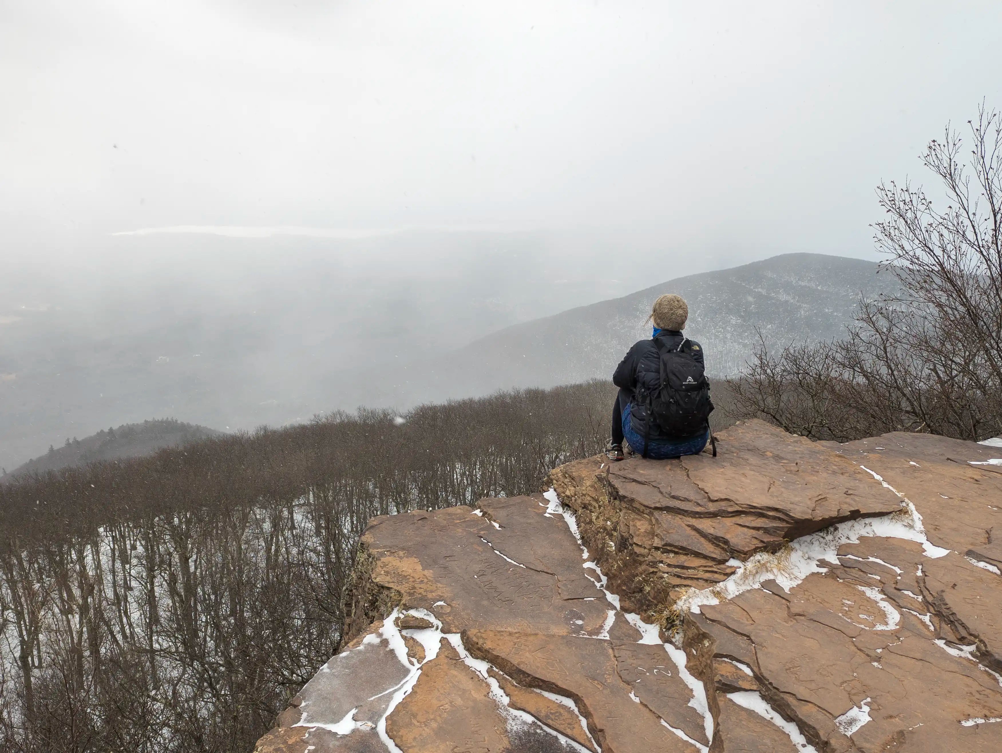 A female hiker siting on a rock in the snow at an overlook on Overlook Mountain in New York wearing the Ancash Packable Daypack by Deep Ascent.