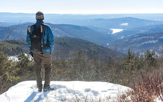 A male hiker standing on a viewpoint in the snow overlooking the Catskill Mountains in New York wearing the Ancash Packable Daypack by Deep Ascent.