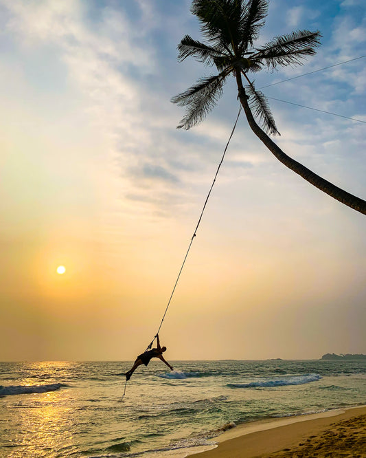 A woman swinging on a rope from a palm tree at sunset at Dalawella Beach, Sri Lanka.