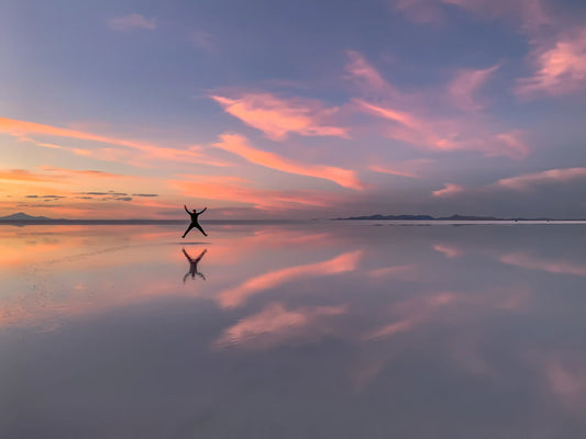 Silhouette of a person leaping in the air over the mirror-like Uyuni Salt Flats in Bolivia, with a vibrant pink and purple sunset reflected in the water.